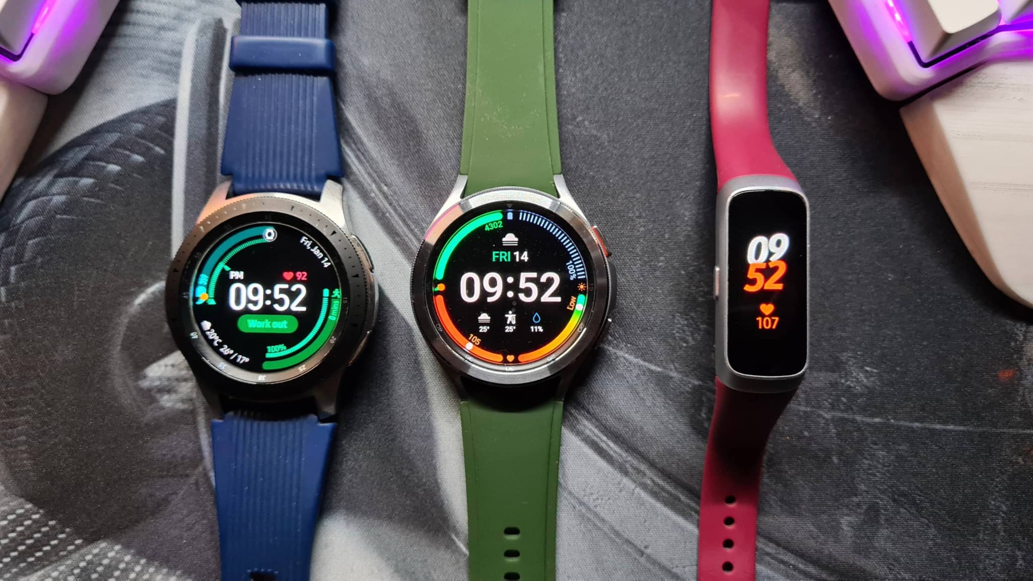 The image displays three smartwatches with their straps, arranged vertically side by side. Each watch has a colorful strap: one is blue, one is pink, and one is green. They all have digital screens displaying various functions and colors, including the time, date, and potentially fitness tracking or app notifications. The watches seem to be of different models or brands, based on their design and screen layouts. They are presented on a surface that could be a table or countertop with an indistinct background, and no additional context or items are visible in the image.