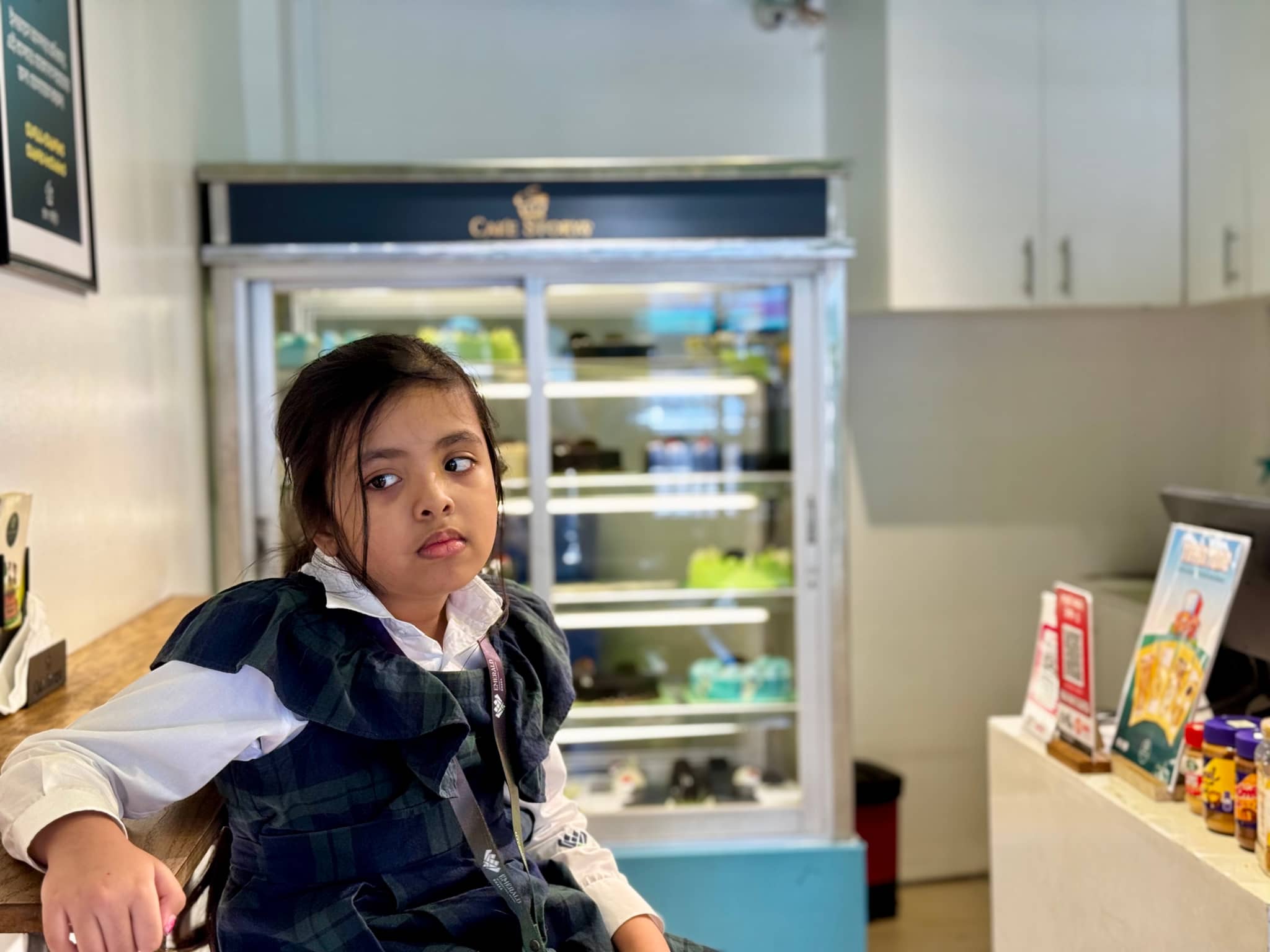 The image depicts an indoor setting, likely a café or store with a prominent counter area. Two individuals are seated at the counter: a child to the left and a woman to the right. Both appear to be engaged in conversation or observation of items on display, such as food in containers or shelves stocked with various products. The environment includes lighting fixtures, a glass panel that suggests a transparent separation between the counter area and the rest of the space, and wall-mounted signs or advertisements.