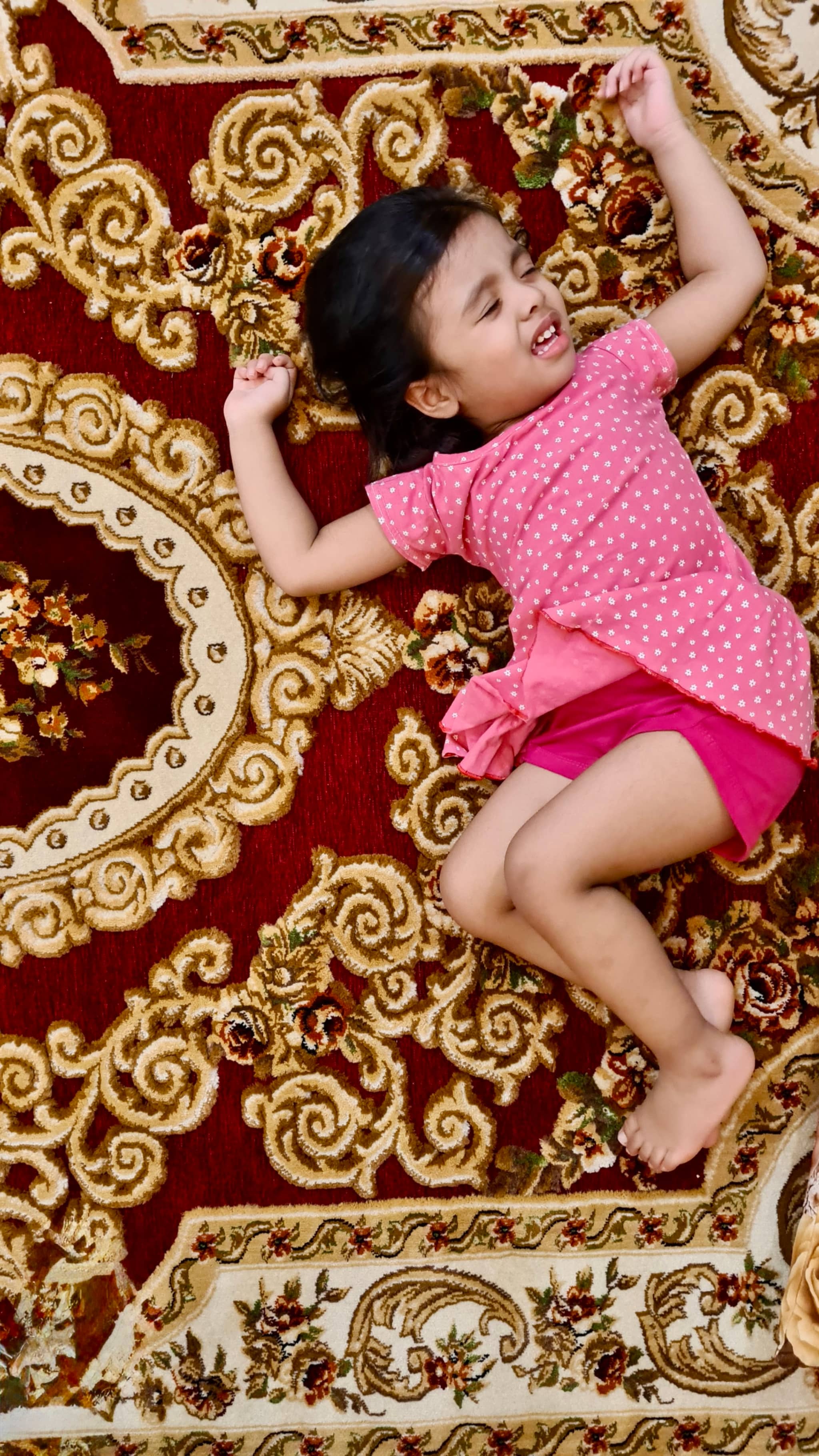 The image depicts a young child, who appears to be a girl, lying on her back with her arms spread out as if she's just fallen or in mid-play. She is wearing a pink dress and has dark hair. The child is on a surface adorned with ornate, intricate patterns that resemble traditional Indian designs. The background is indistinct due to the focus on the child.