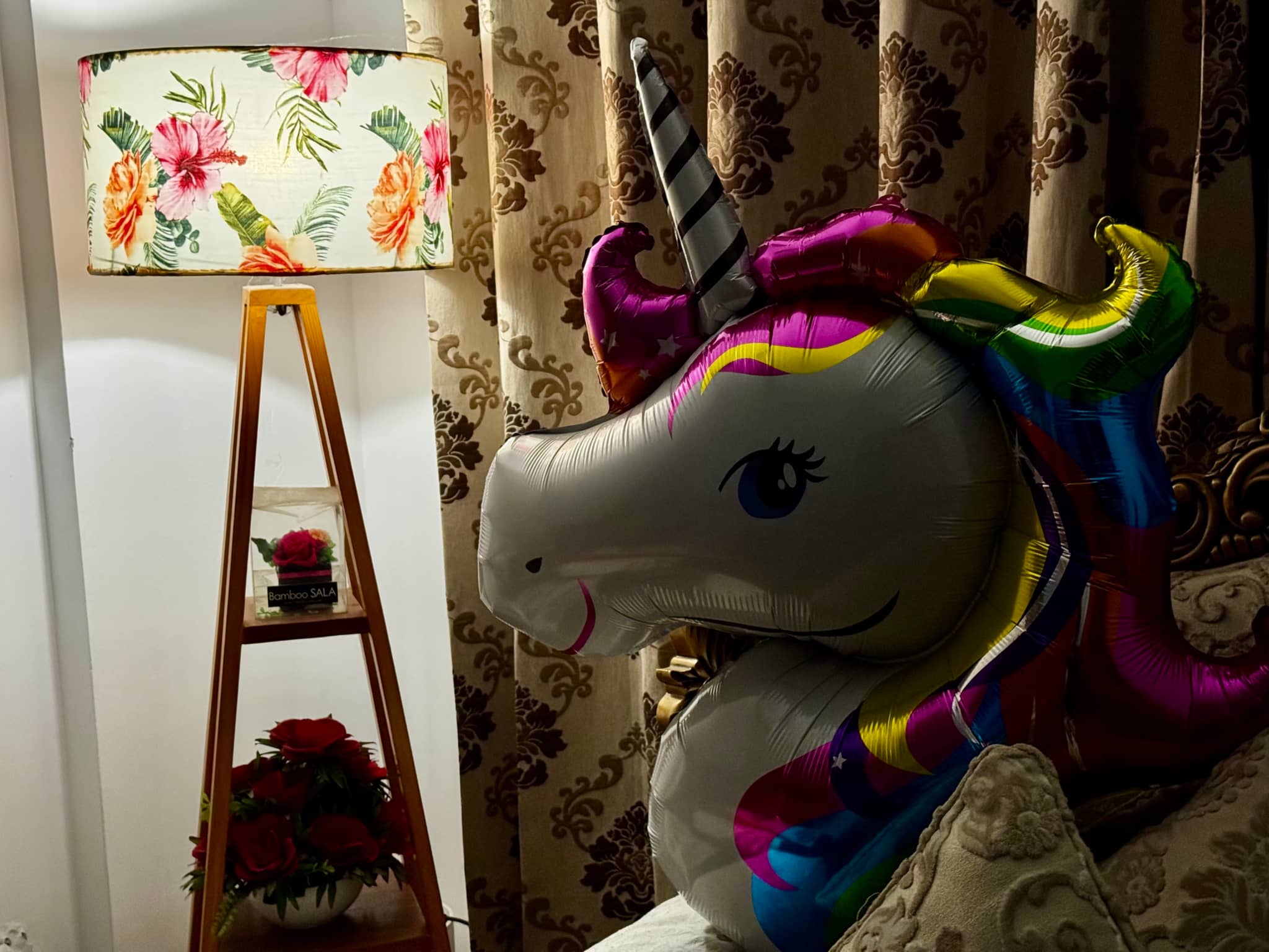 The image features a colorful toy unicorn sitting on a surface, placed next to a white wall. The unicorn has a large mane and tail, with different shades of pink and purple. It stands upright, with one leg elevated. Behind the unicorn, there is a framed picture or artwork hanging on the wall. Next to this frame is a tall wooden ladder leaning against it. To the left of the unicorn, a lamp with a floral shade casts light onto part of the unicorn's body. The background has a patterned fabric and a portion of a window can be seen.