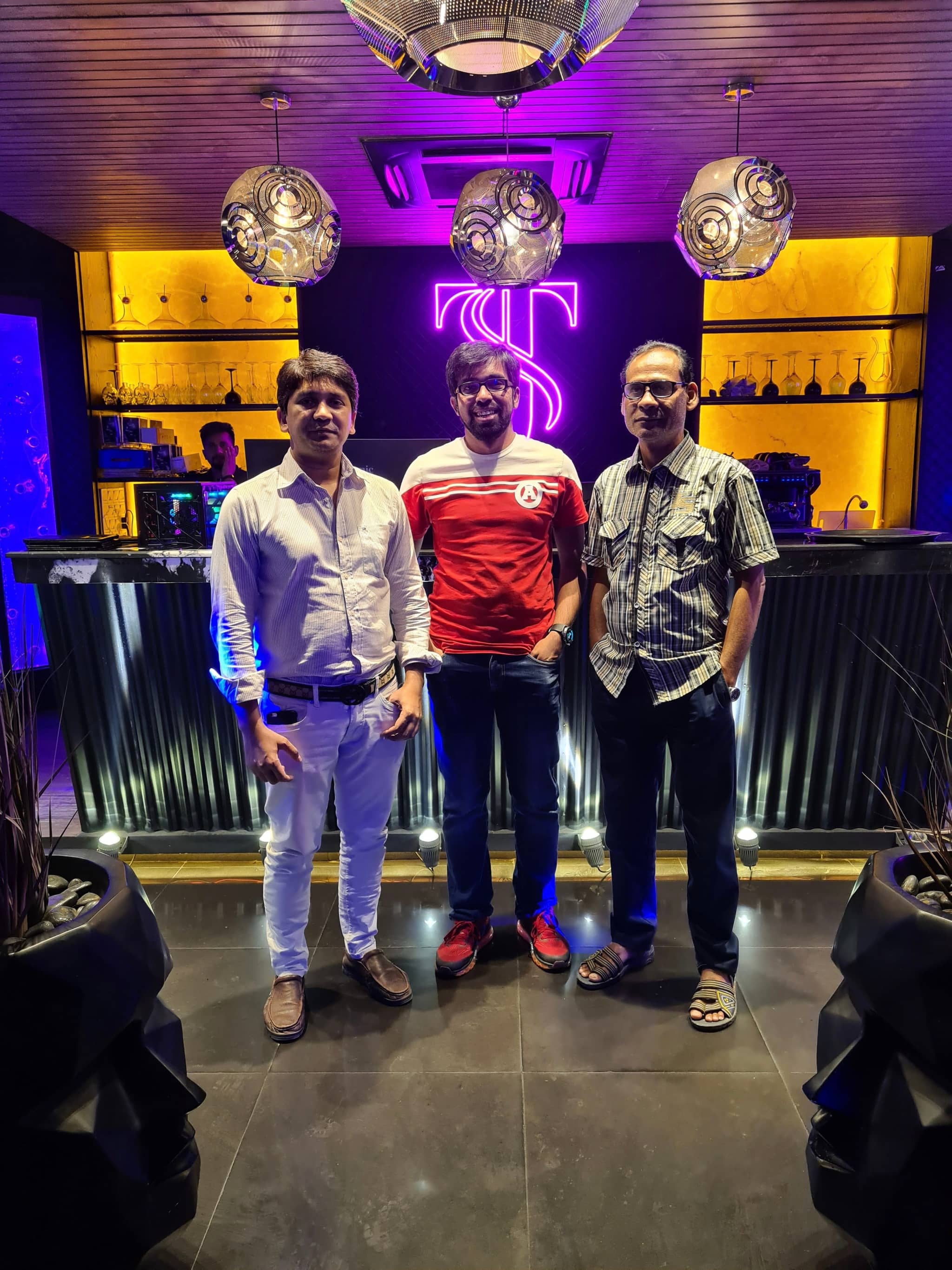 The image shows three individuals standing in front of a bar with a lively atmosphere. They are posing for the photo, with two men flanking another man who is holding an item that resembles a trophy. All three appear to be enjoying themselves at a social event. Behind them, the bar is adorned with decorative elements and has a cozy ambiance.