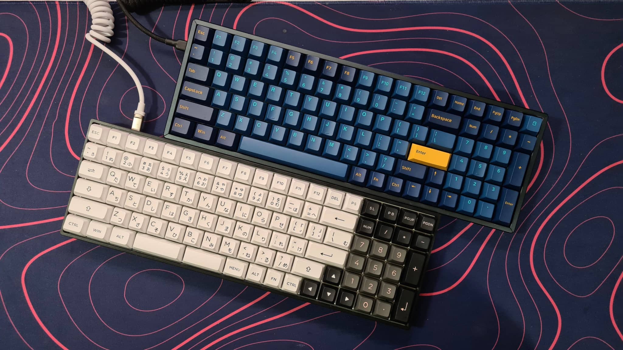 The image displays two computer keyboards placed side by side on a surface with a geometric pattern. The keyboard in the foreground has a black body with white keys, while the one behind it appears to have a blue and yellow color scheme. Both keyboards are shown with the numeric keypads facing the viewer, and the space bars are visible. In the background, there is a hint of a computer mouse, suggesting this might be a workspace or a store specializing in computer peripherals. The style of the image seems to be an informal photograph meant to showcase the keyboards.