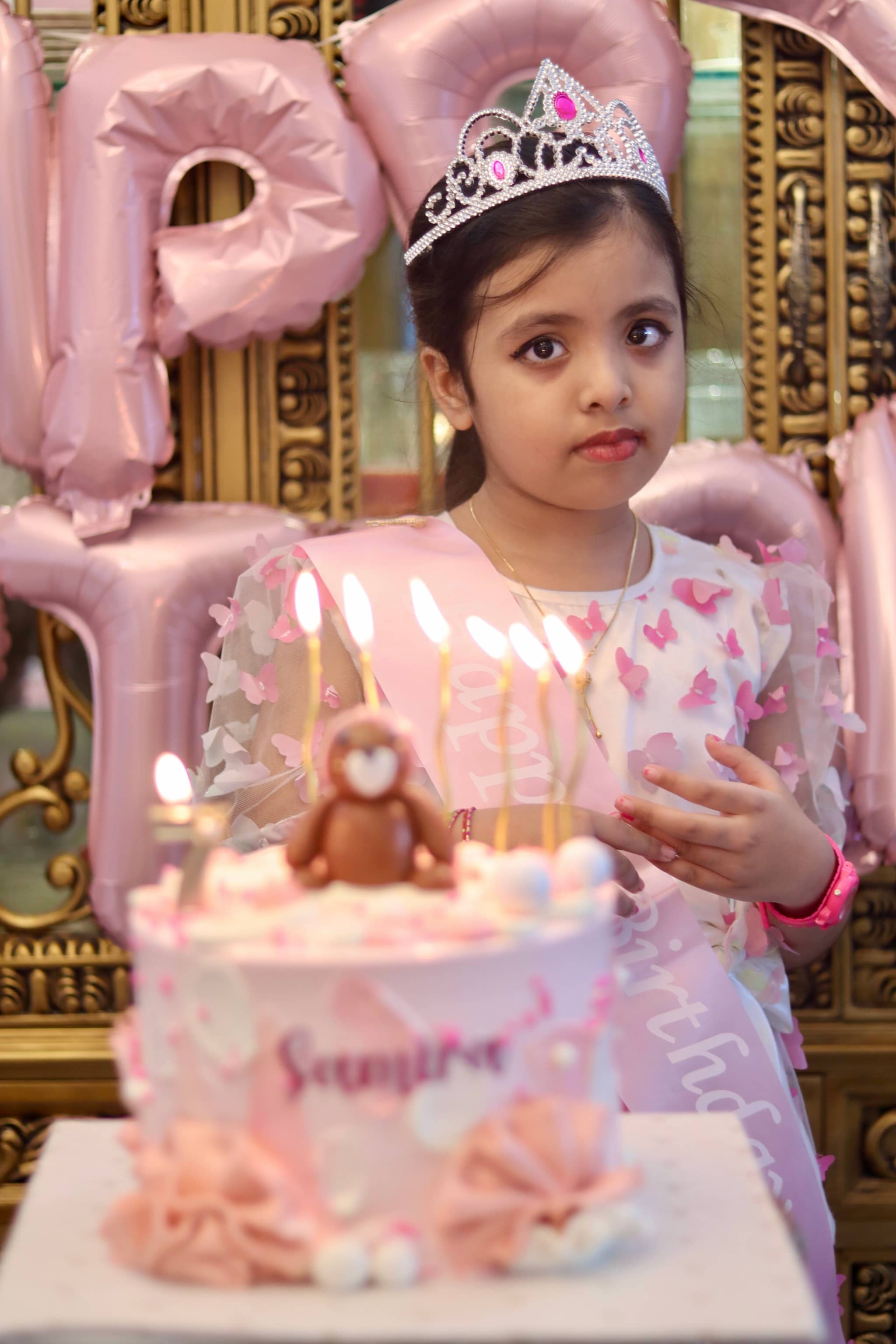 The image captures a young girl, possibly a princess given her attire. She is seated in front of a birthday cake with candles, suggesting a celebration of some sort. There is a pink balloon in the background, and a large cardboard sign that reads "Happy Birthday." It appears to be a joyful birthday party event, likely for a young girl celebrating her special day.