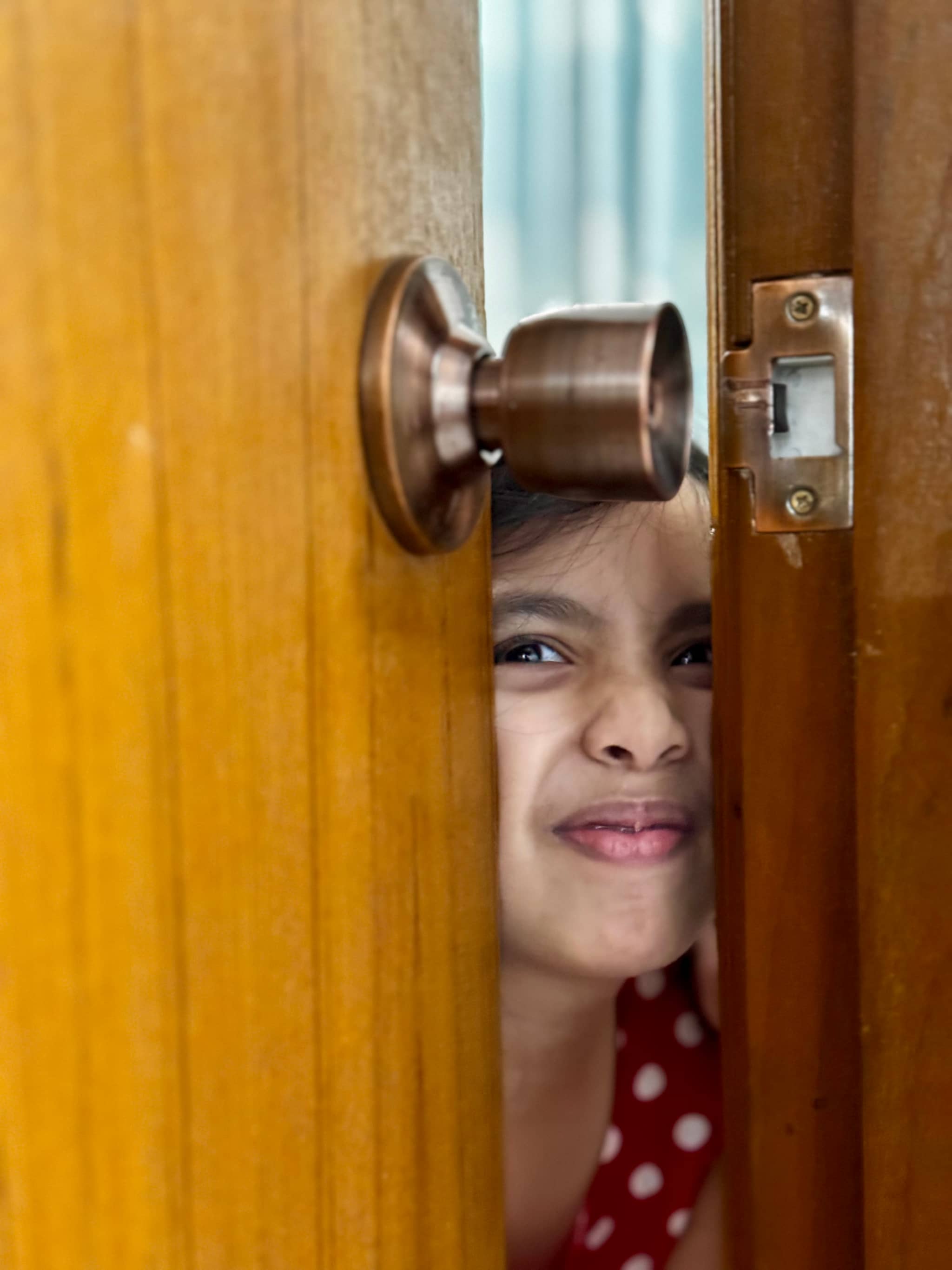 In the image, there's a person standing in front of a wooden door, looking through the door with a curious and surprised expression. The door has a brown finish and a bronze doorknob. The individual appears to be a young child or a young adult, dressed in casual clothing, peering out from behind the door, which is open a little bit at the top.
