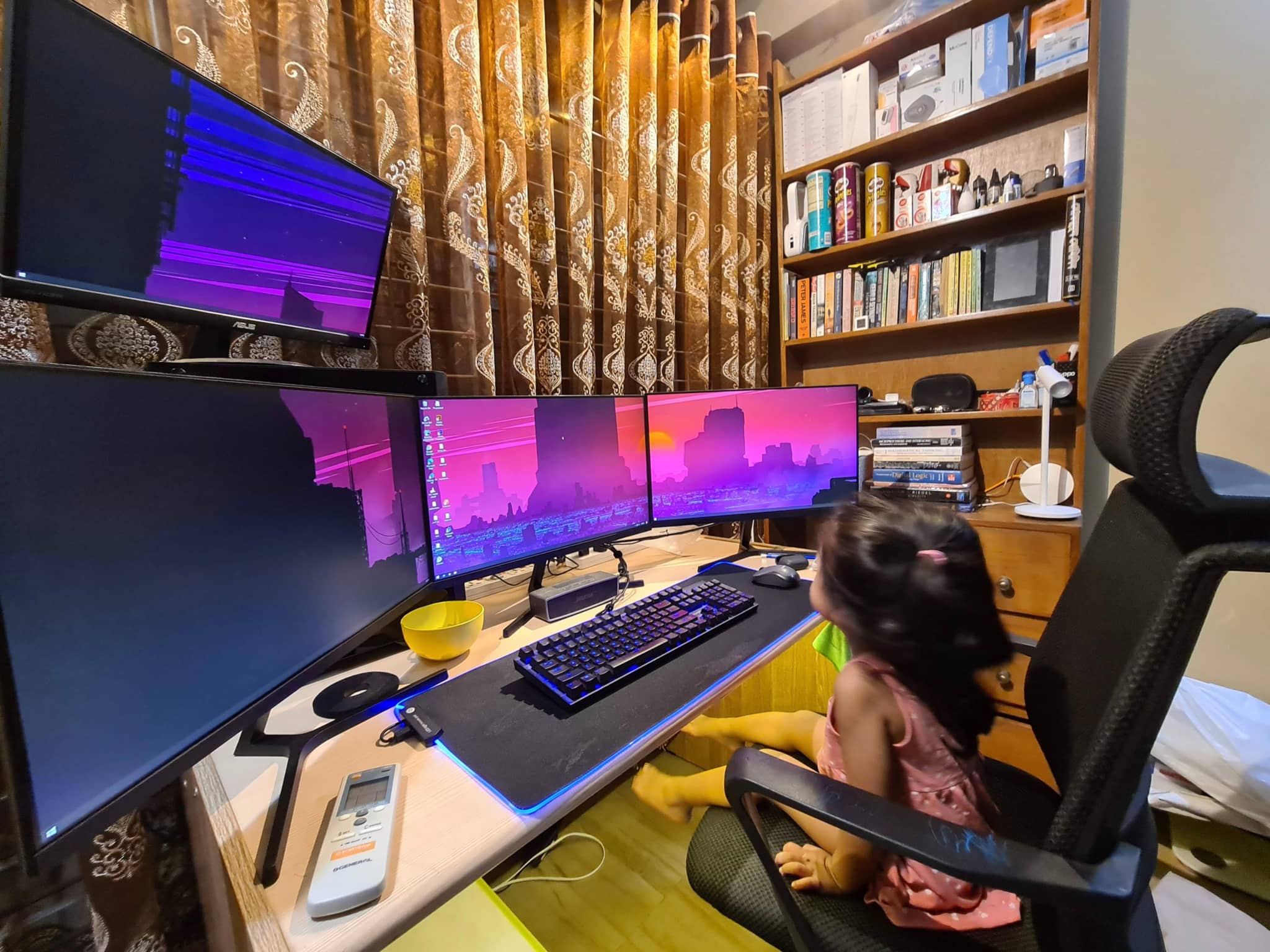 The image depicts a home office setting. There's a large, multi-monitor computer setup with various screens and peripherals, including a keyboard, mouse, and headset. A person is seated at the desk, using one of the monitors; their back is turned to the camera. In front of the screens, there are two chairs: one with a human figure partially visible from behind, and another with no one sitting in it. The room has a cozy ambiance with bookshelves on either side, providing a personal touch. The overall scene suggests a space dedicated to work or gaming, with a comfortable and functional environment.