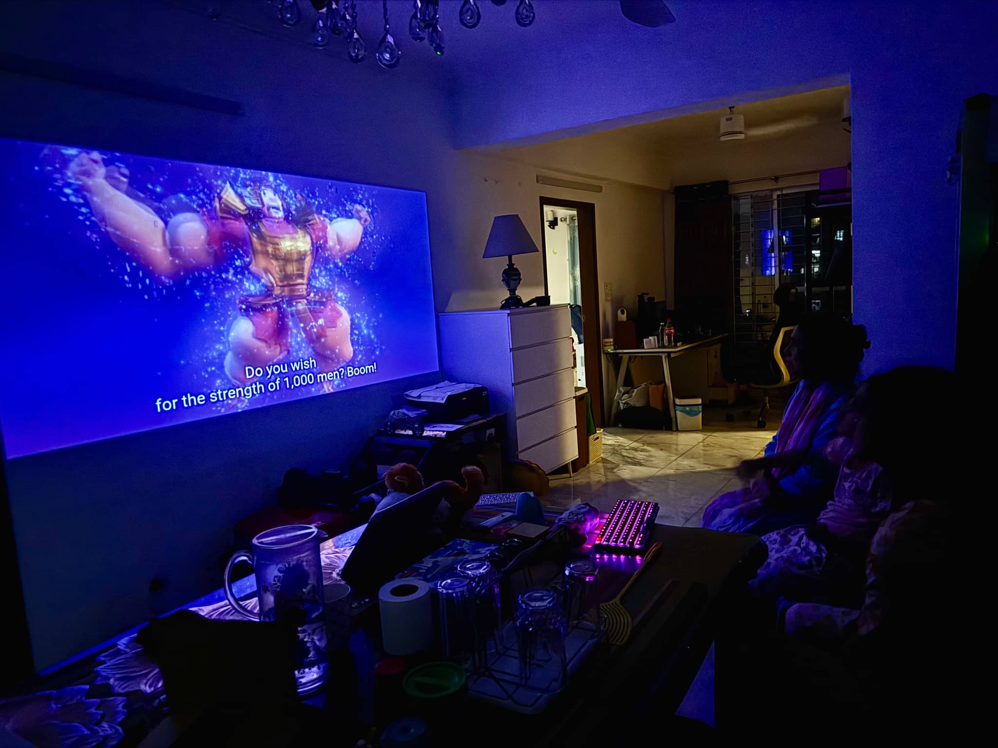 The image depicts an indoor living space at night, where a large projection screen displays colorful graphics that appear to be related to video gaming or animation. The room is dimly lit, emphasizing the bright, blue-tinged images on the screen. In the background, there are objects indicative of a lived-in area, such as furniture and decorations, but they are not clearly visible due to the focus on the projection screen.
