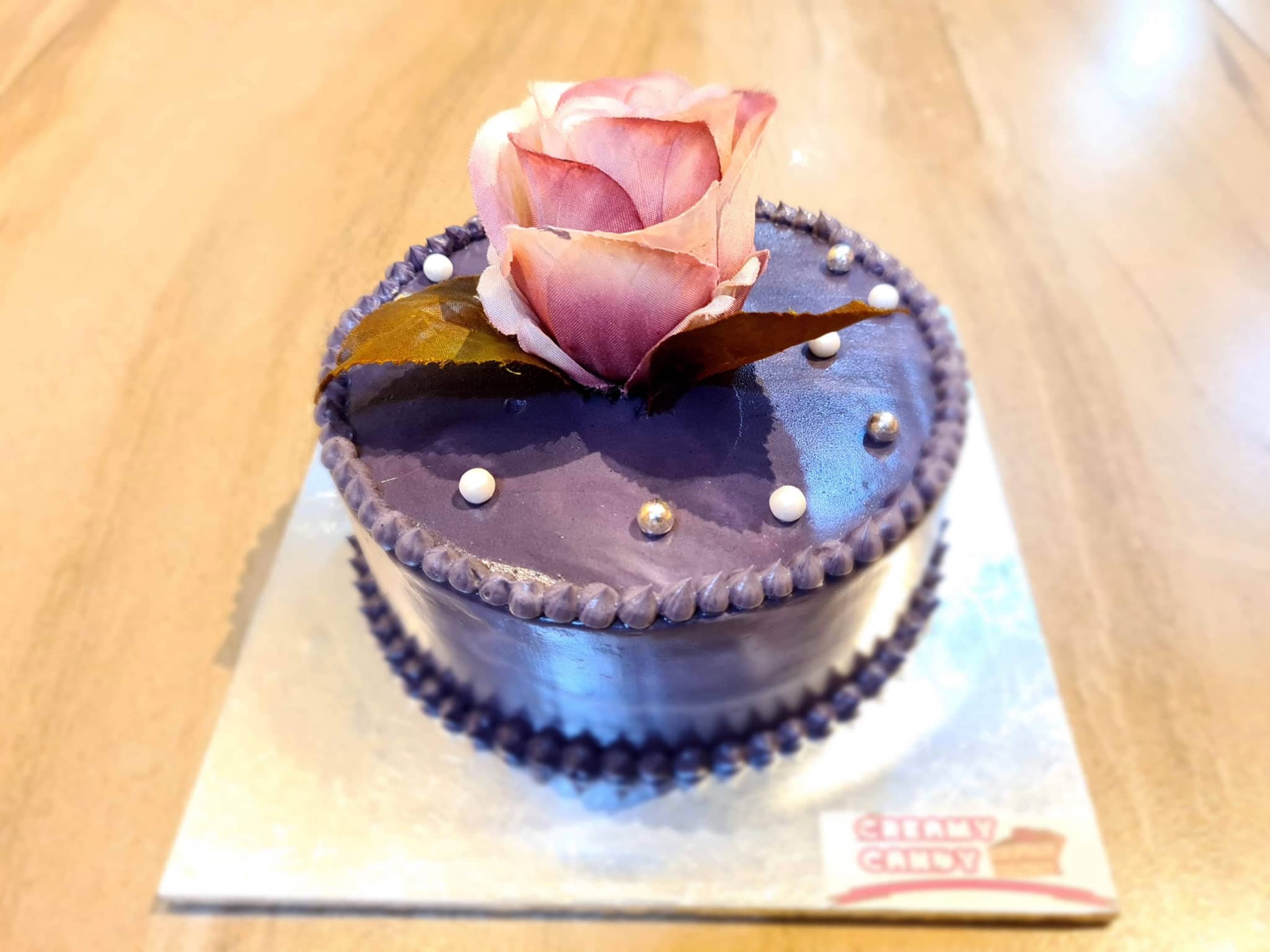 The image features a small purple cake with a single rose on top, sitting on a white surface. The cake has a design that includes what appears to be pearls and possibly some icing decorations, but it's not entirely clear from this angle. The background is blurred, highlighting the cake as the main focus of the image.