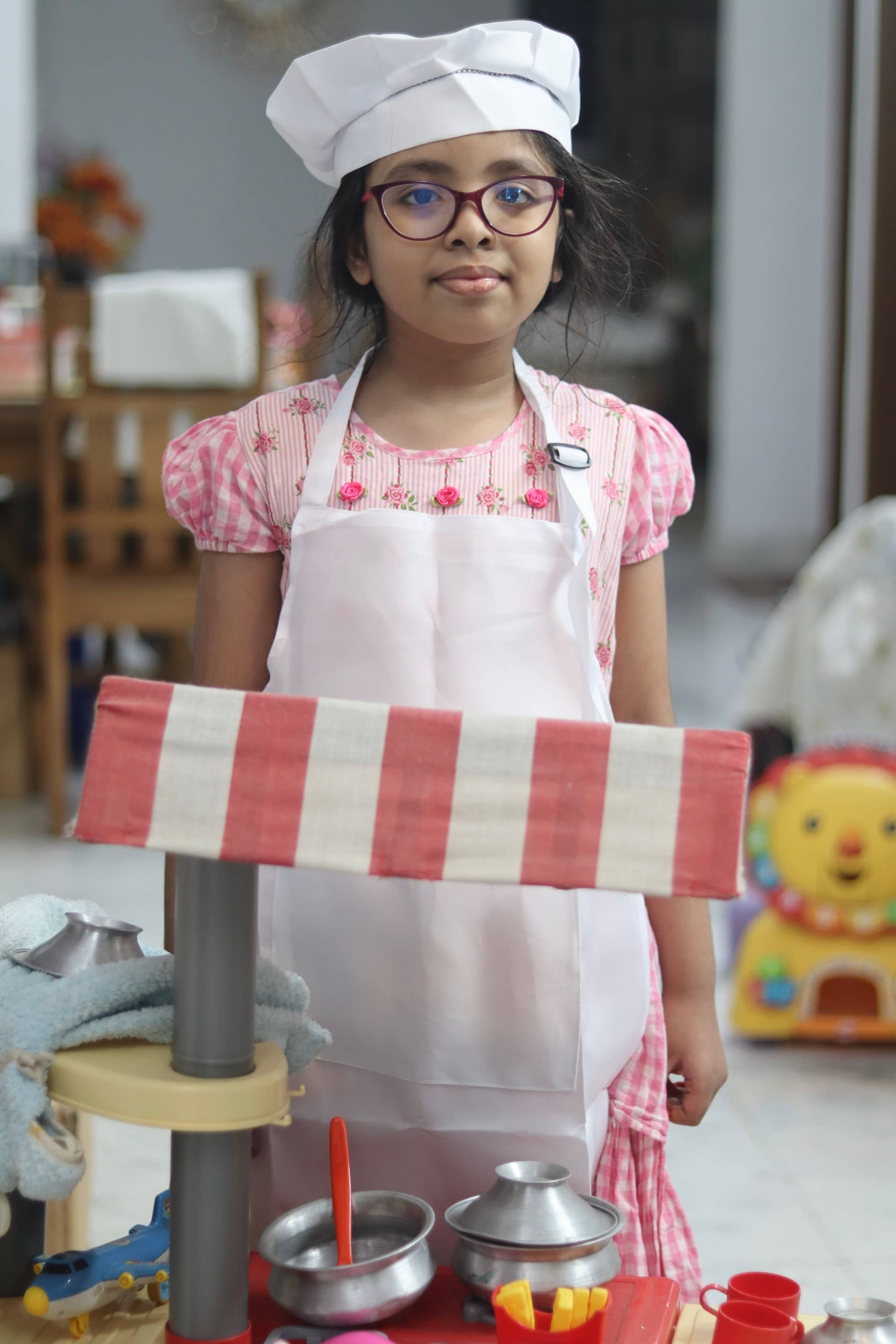 This is a photograph of a young child dressed in a chef's outfit, holding a fake red and white checkered hat, positioned in front of a table with various kitchen items. The setting appears to be indoors, possibly a home, with a tiled wall in the background.