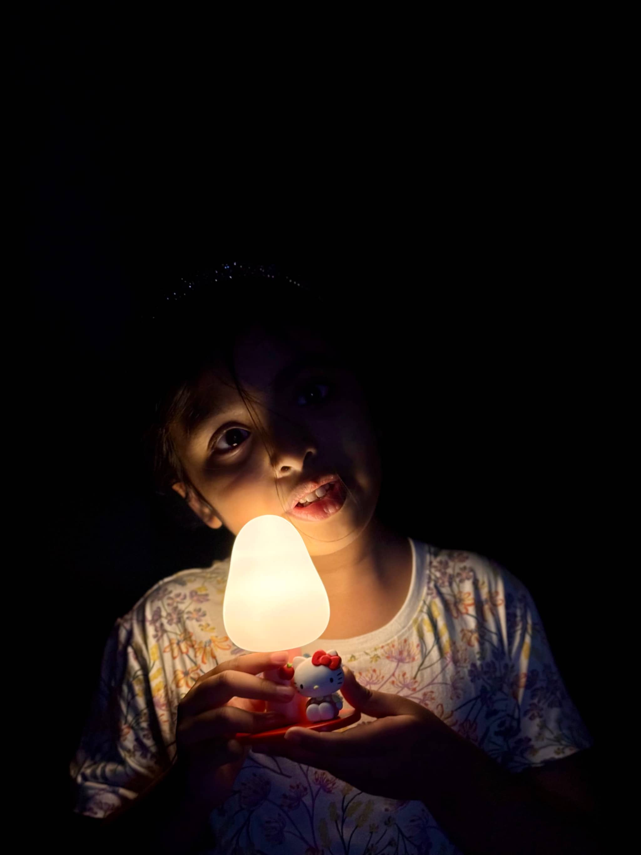 The image depicts a person holding a lit candle in front of a window. The individual appears to be a young girl with dark hair, and she's looking towards something outside of the frame. She's holding the candle in her right hand, which emits a warm glow.