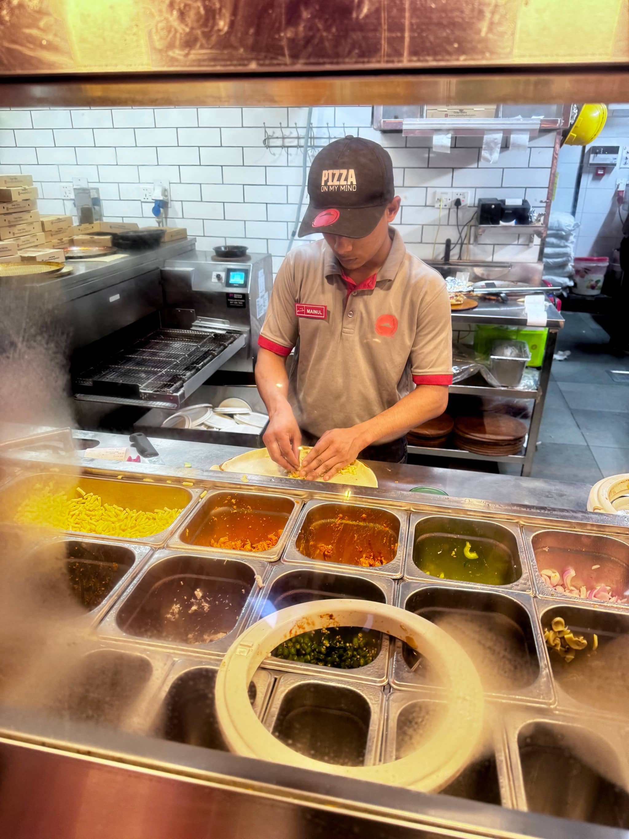 This image depicts a restaurant kitchen scene. A chef, wearing a white apron and a black cap with red lettering, is working at a counter in front of open food containers and various condiments. The chef appears to be preparing food, focusing on the task at hand. In the background, there are additional counter spaces equipped with more cooking tools and condiments. The image captures the bustling atmosphere of a commercial kitchen.