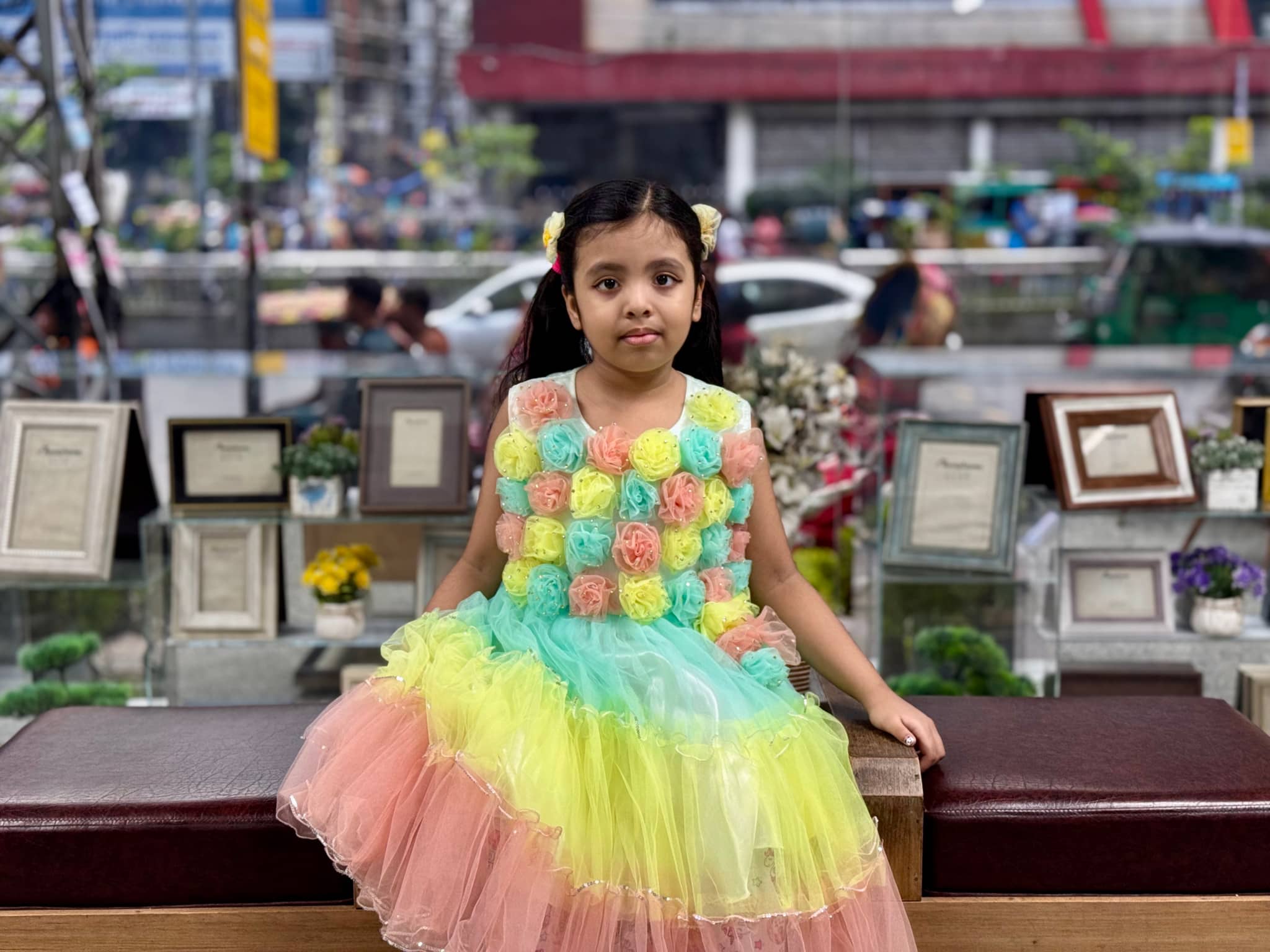 The image features a young girl with a colorful dress, posing in front of a display that includes various items on shelves. She appears to be sitting at a bench and has a sweet and joyful expression. Behind her, there seems to be an indoor environment with framed pictures on the wall. The setting suggests this could be a public space like a museum or a gift shop.