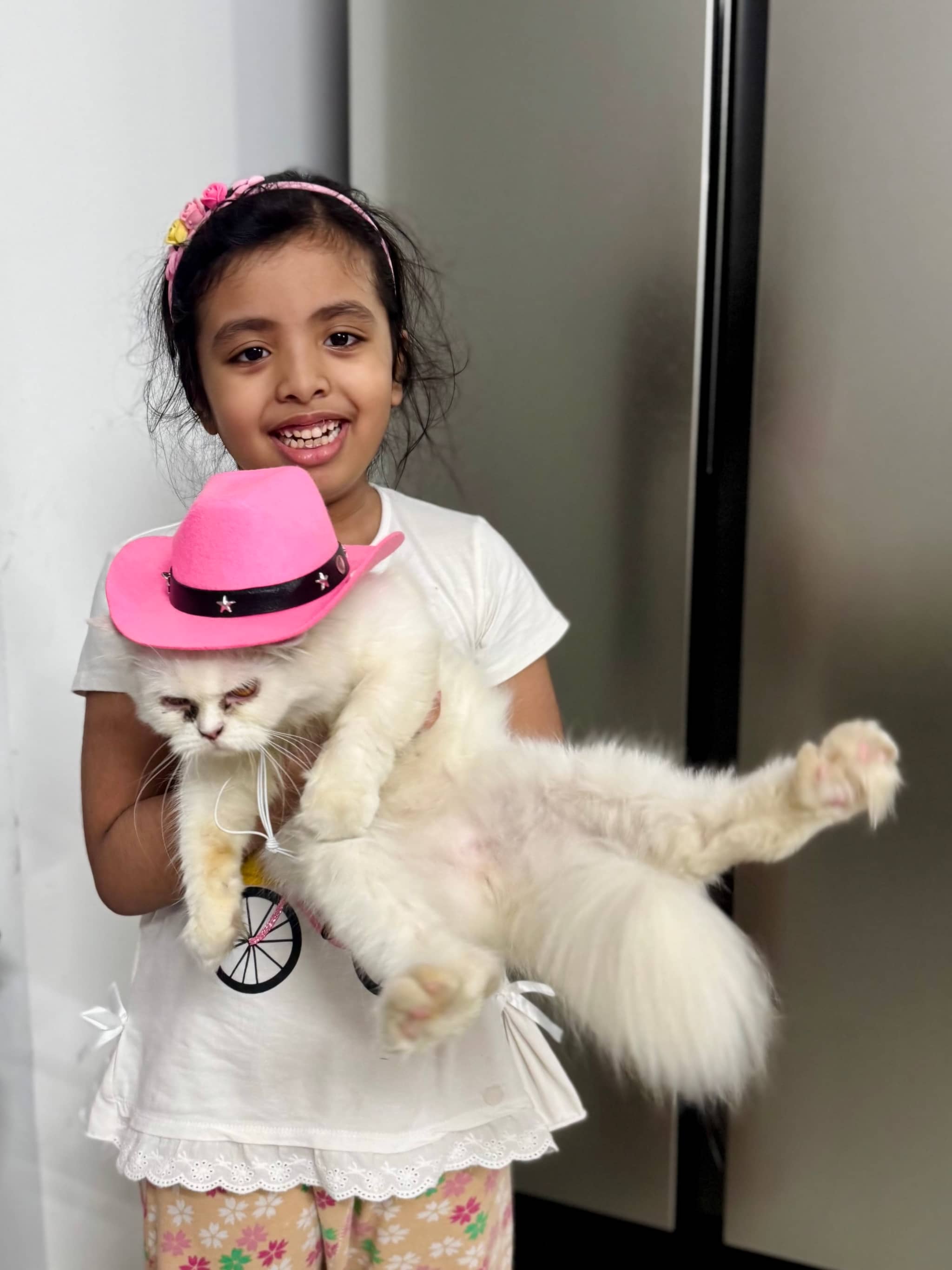 In the image, a young girl is standing next to a cat. The cat is wearing a pink hat. The girl appears to be holding the cat, who seems to be comfortable with her touch. She is dressed in casual attire and looks happy to have her pet cat with her.
