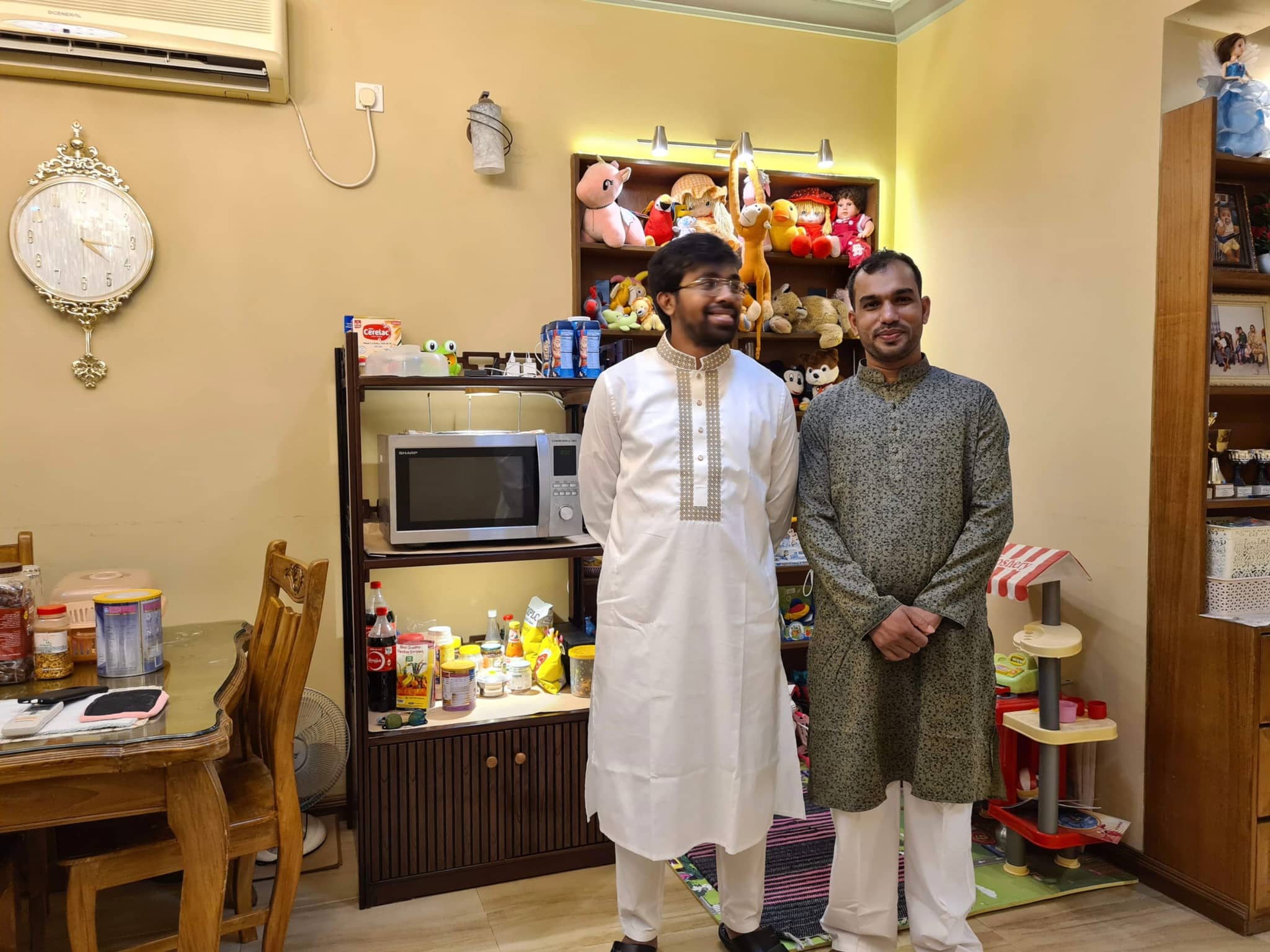 The image depicts two men standing together in an indoor setting that appears to be a kitchen or dining area. The man on the left is wearing traditional Indian attire, consisting of a kurta and a sherwani, with a white turban on his head, while the man on the right is dressed in Western casual wear. Both are smiling and seem to be posing for the photo. Behind them, there's a wall-mounted clock and some decorative items. The kitchen counter has various items on it, such as food packaging and a small plant. The room has a warm ambiance with lighting that casts soft shadows.