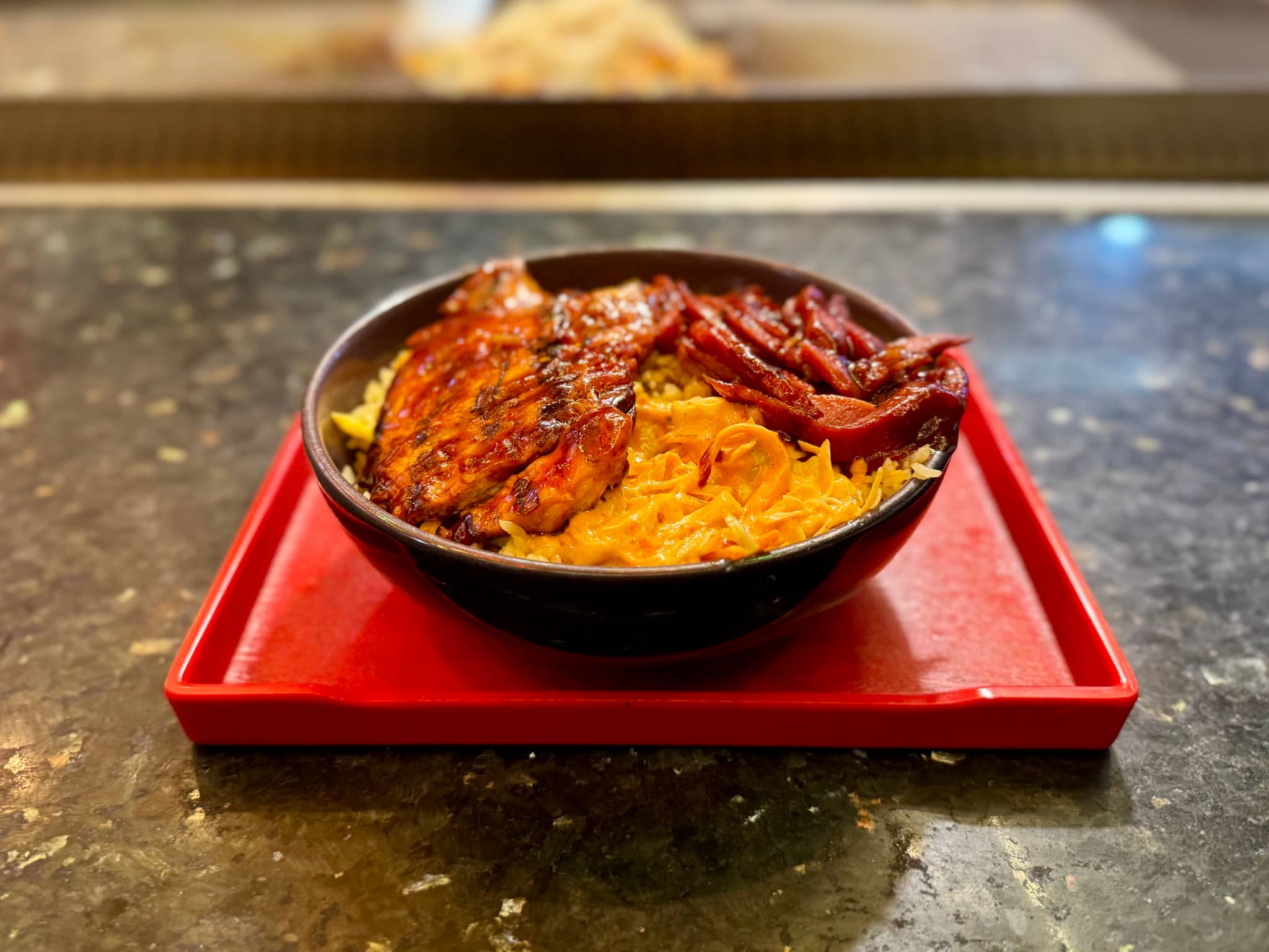 The image features an oriental meal served on a red tray atop a countertop. There are two bowls: one with what appears to be steamed rice and another with slices of grilled meat, possibly chicken. Beside the dishes are a small bowl filled with a sauce that could be chili or curry, and a portion of fried noodles in a separate bowl. The tabletop has a dark surface, and the overall setting suggests a casual dining environment. There is also a blurred image of what looks like another food item in the background, possibly part of the same meal or from a nearby station.