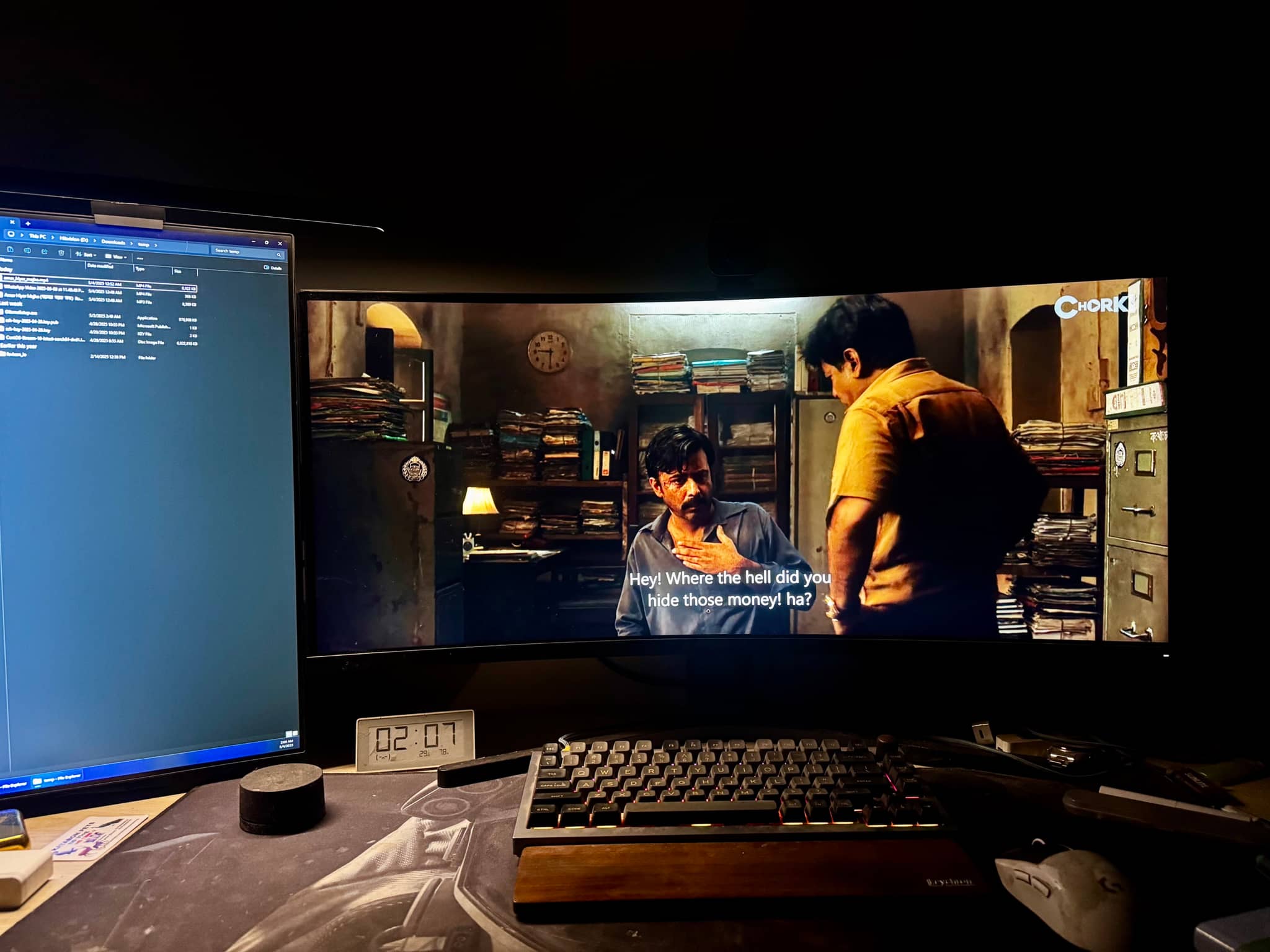 The image depicts a home theater setup with a large widescreen monitor prominently displaying a movie scene. Two individuals are visible in the scene, engaged in what appears to be an intense conversation. There's a well-lit room, with a gaming computer partially visible on the left side of the frame.