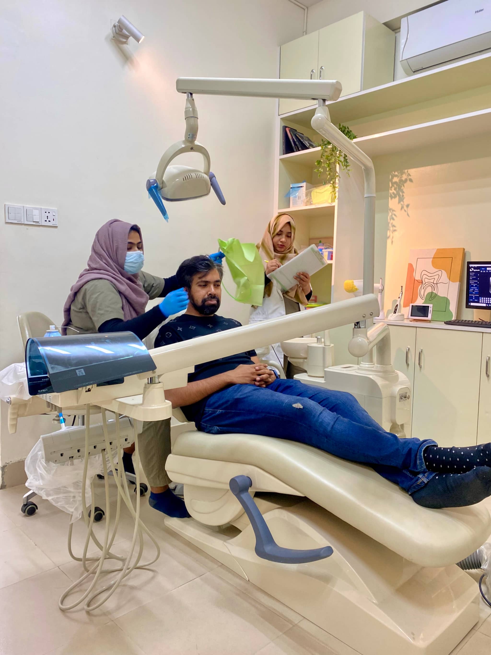 The image depicts an indoor dental practice setting. In the foreground, there's a person seated in a dental chair, seemingly waiting or preparing for a dental procedure. To the left, there are two individuals who appear to be dentists or dental hygienists, standing and observing the seated person. They are wearing face masks and stethoscope necklaces, indicative of their profession.

The practice room has a modern look with a white tiled floor and walls painted in light colors. On the right side of the image, there is dental equipment visible, including what appears to be a dental chair mounted on an adjustable arm for different angles of work during procedures. The overall atmosphere suggests a professional and sterile environment, typical of a dental office.