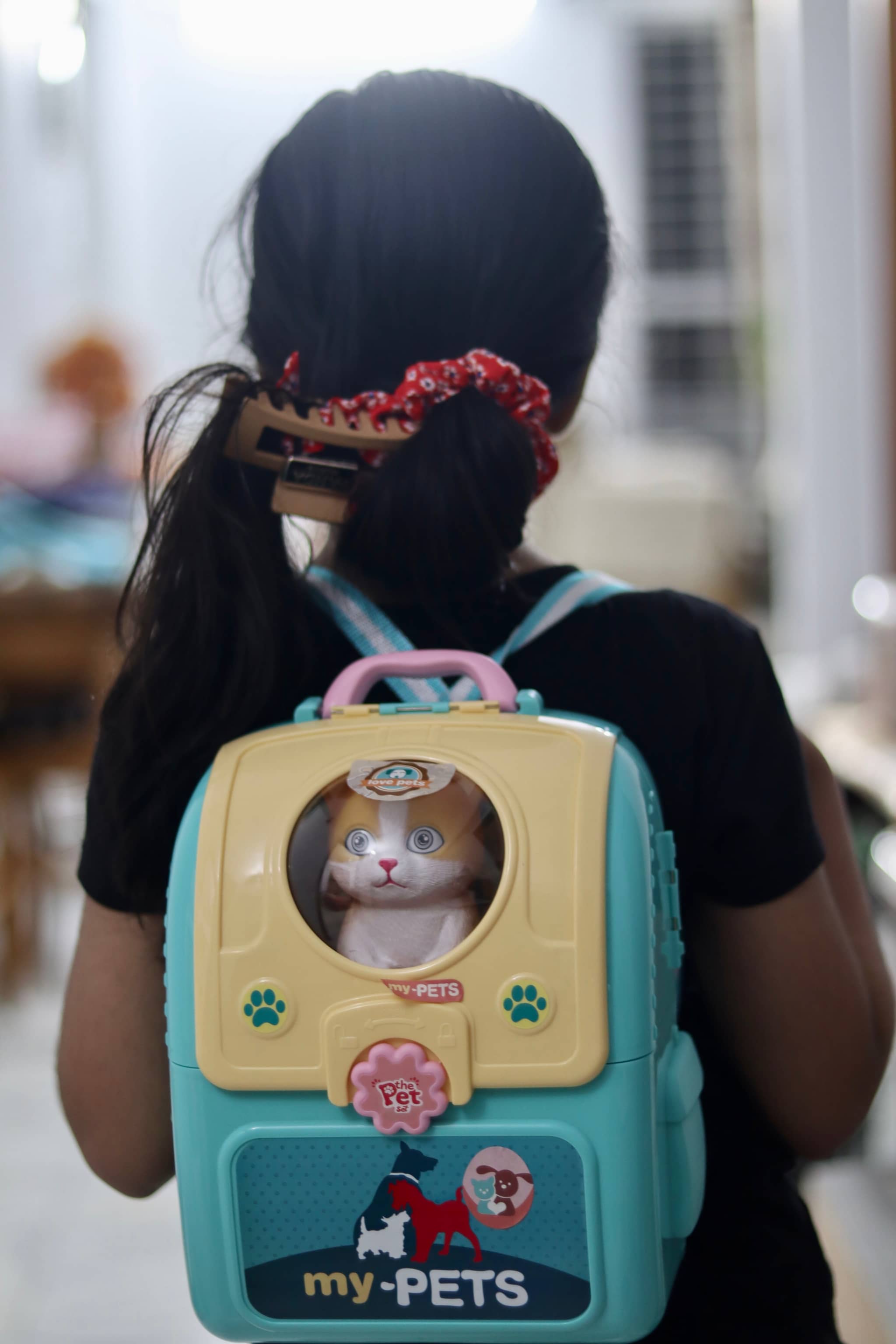 The image depicts a young child, possibly a girl, with dark hair and a braid. She is wearing a black shirt and has a pink backpack on her back. On the backpack, there is an image of a cat. In front of her, there appears to be a watermark or logo that reads "MY PETS" with the words "PETS" and "CATS" below it. The child is standing in a room with indoor lighting visible from above.