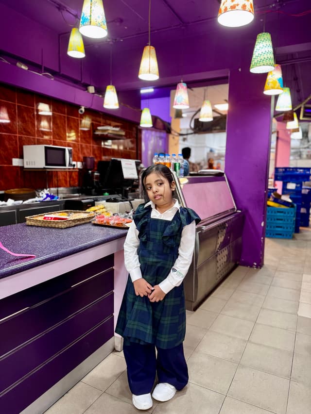 In the image, there's a young girl standing in front of a counter. The counter has a variety of pastries and cakes on display. Behind her, there are shelves with more items for sale. On either side of her, there are decorative lights hanging from the ceiling. To the right, there is a small part of what appears to be an establishment with a purple-themed interior. The girl seems to be posing for the photo with a slight smile on her face.