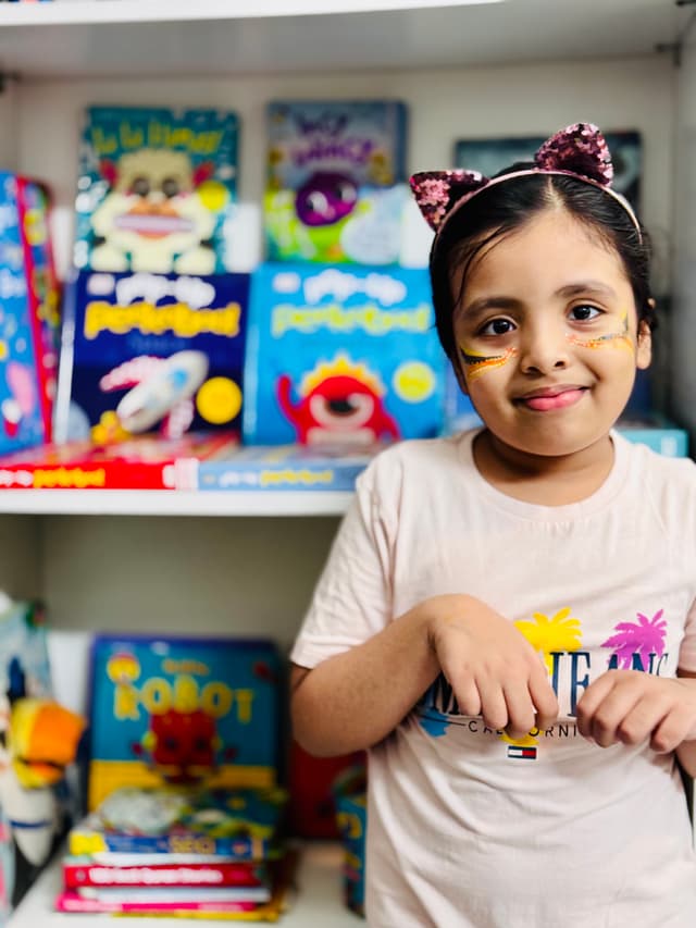 This is an image of a young girl standing in front of a collection of colorful children's books. She has a playful expression, wearing a white top with cat ears on her head. The setting appears to be a room dedicated to children's literature or learning, as evidenced by the variety of books and toys behind her.