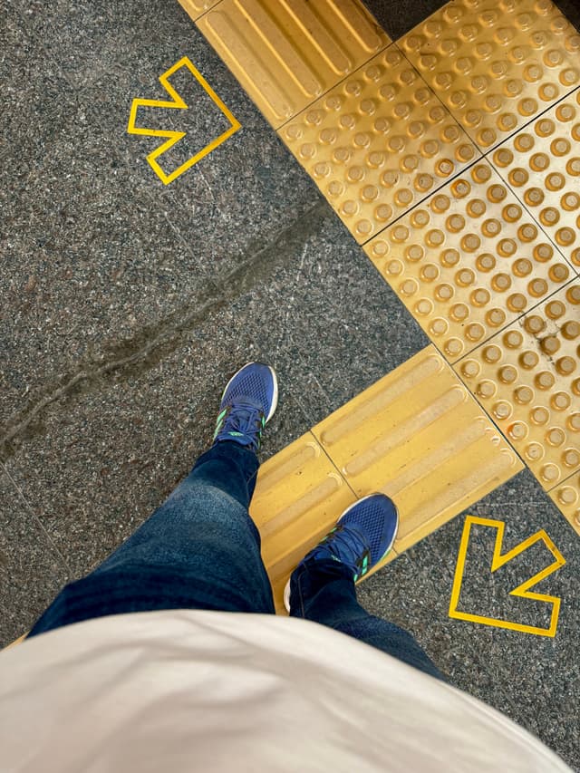 The image features a person standing on an escalator at what appears to be a subway station. The individual is wearing a white t-shirt, blue jeans, and athletic shoes with a checkmark design. They are also wearing shorts. The escalator has yellow lines painted on the right side, indicating directions for passengers. At the top of the image, there are two arrows pointing to different directions, suggesting that the person is at a junction or change of direction in the station's layout.