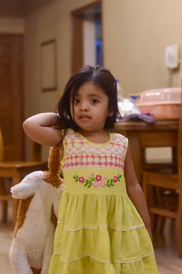 The image shows a young girl with dark hair, wearing a yellow and green dress. She is standing in a room that appears to be a living area, as there is a dining table in the background. Her gaze is directed downwards, away from the camera.