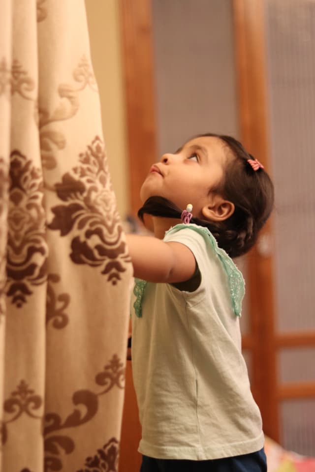 The image depicts a young child standing in front of an open window with curtains, looking out. The child appears to be focused on something outside the frame. The environment suggests a residential interior with wooden floors and walls.