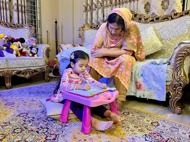 The image depicts a room where a woman and a young girl are present. The woman appears to be sitting on a couch and has her attention directed towards the child, who is seated on the floor with a tablet or small laptop in front of them. The child seems to be using the device. Both individuals seem to be engaged in an activity or conversation related to the screen content. They are indoors, with decorative elements such as cushions and curtains visible.