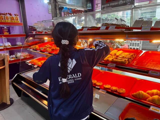 The image depicts a person, presumably a child, standing in front of a shop counter that displays an assortment of food items on shelves. The environment suggests it's inside a store or market. The child appears to be observing the products, possibly contemplating a purchase or just being curious. The setting has a casual atmosphere with natural light illuminating the interior.
