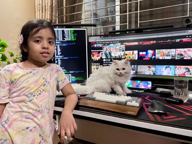 The image features a young girl standing next to a cat at a computer desk, posing in front of a desktop monitor that displays a variety of windows, including what appears to be video editing software and web browsers with multiple tabs open. To the right, there's an adult female seated, also at the desk with the cat on her lap. The room is well-lit and has decorative elements such as a vase, books, and a keyboard in the foreground.