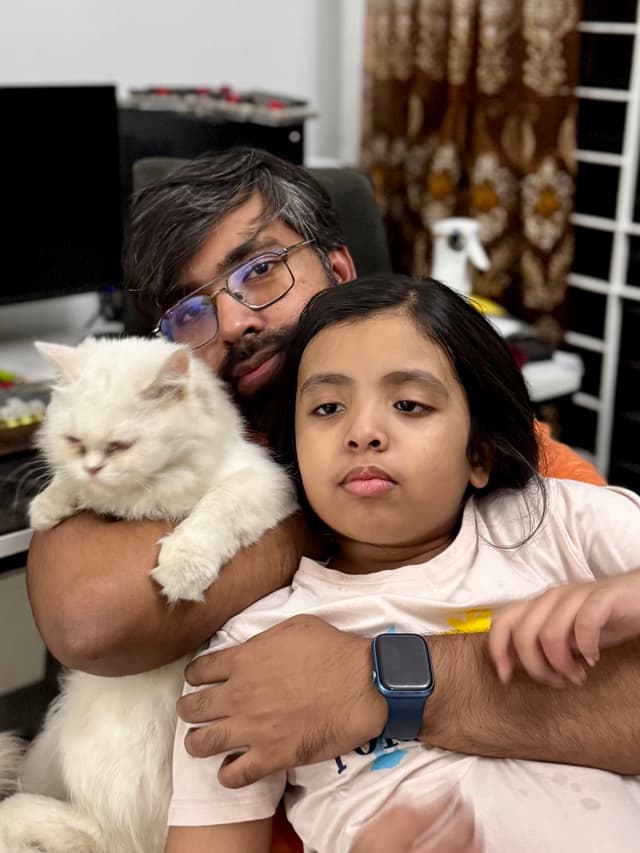 The image depicts a person sitting at a desk with their arm around another individual who is holding a cat. They are all smiling and looking towards something outside of the frame.