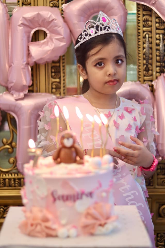 The image captures a young girl, possibly a princess given her attire. She is seated in front of a birthday cake with candles, suggesting a celebration of some sort. There is a pink balloon in the background, and a large cardboard sign that reads "Happy Birthday." It appears to be a joyful birthday party event, likely for a young girl celebrating her special day.