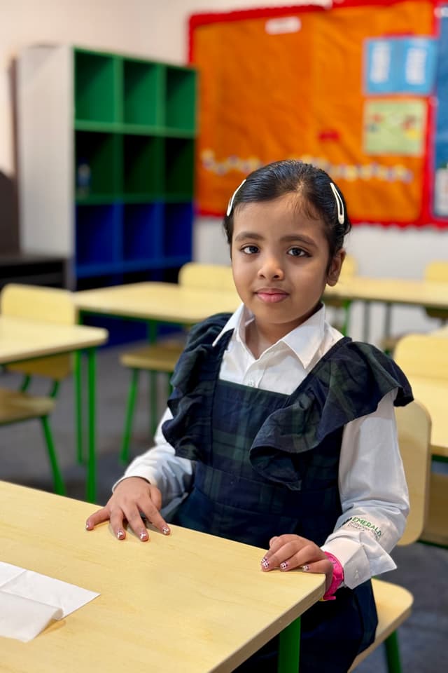 The image depicts a young girl sitting at a desk in a classroom setting. She appears to be a student, as suggested by her attire which includes a school uniform. Her facial expression is neutral, and she is looking directly at the camera. The room has a simple setup with desks and chairs, typical of an educational environment. There is a bookshelf behind the girl, and a flag hanging on the wall above her head, though its details are not clearly visible in this image. The overall atmosphere seems to be calm and orderly, indicative of a school or similar educational establishment.