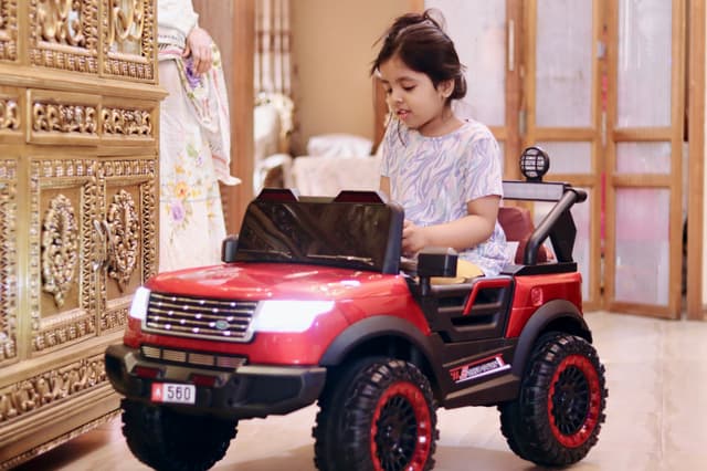 The image features a young child sitting in a toy jeep, which appears to be an electric vehicle with red accents. The toy car has large black wheels and a design that mimics a real SUV, including a hood scoop and front grille. It's painted in a vibrant combination of red, white, and black colors, adding to its realistic look.

The child is wearing casual clothing, with one arm visible as they seem to be enjoying the toy ride. The setting looks like an indoor room with a decorative ceiling and what appears to be a traditional wooden door or window in the background. The overall atmosphere of the image is playful and cheerful, capturing a moment of childhood fun.