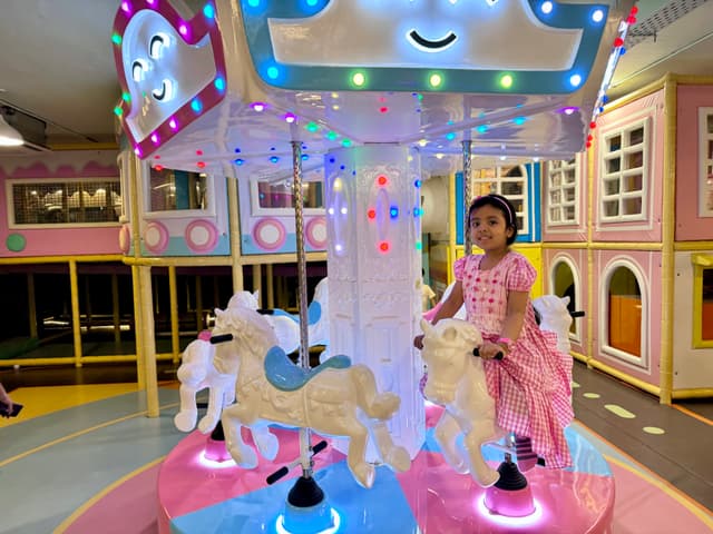 This image depicts an indoor play area with a child standing in front of it, which appears to be designed for small children. The play area includes colorful elements and various structures that seem to promote imaginative play. There are lights illuminating the area, creating a vibrant atmosphere. In the background, there is a structure resembling an ice cream truck or similar cart-like fixture, and above it, a sign with smiley faces. The child seems to be engaged with the play area, possibly posing for a photo.