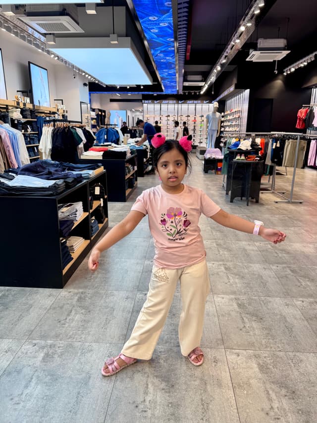 The image shows a young girl standing in an indoor retail space, possibly a store, with shelves and clothing racks in the background. She is wearing a pink top and white pants, with her arms outstretched as if she's dancing or posing for a photo. Her expression seems happy and energetic. There are various signs and logos visible on the storefront glass, including what appears to be a coat brand logo on the right side of the image. The lighting is artificial, suggesting an indoor setting with large windows or doors that allow natural light to enter.