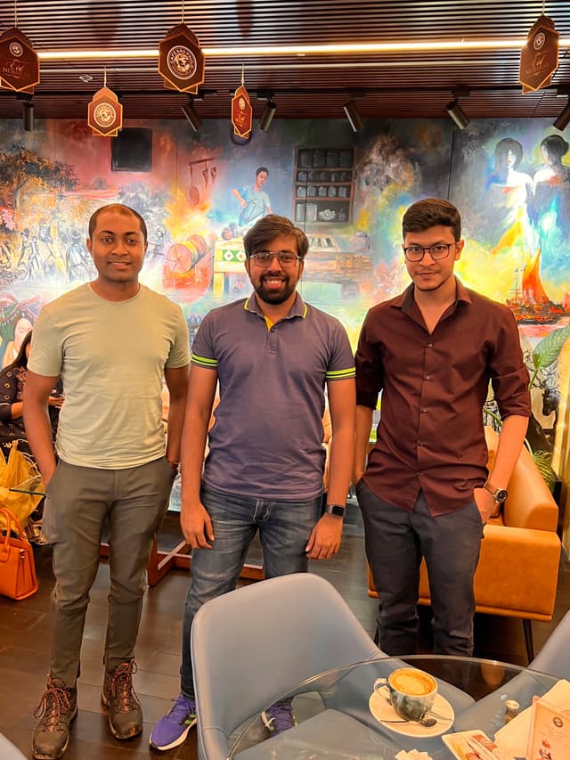The image is a photograph capturing four individuals posing for the camera in what appears to be an indoor setting with a vibrant background, possibly a restaurant interior. They are standing side by side, from left to right: 

1. A person wearing a long-sleeved white top and dark pants, holding what seems to be a menu or flyer in their hand.
2. Another individual wearing a black t-shirt and jeans, standing between the first two people.
3. The third person is wearing a patterned shirt, shorts, and sandals, and has a slight smile on their face.
4. The fourth person on the far right is dressed in a light-colored short-sleeved shirt, dark pants, and sneakers.

All of them are smiling and looking at the camera. The lighting in the room suggests it might be evening or indoors with artificial lighting. There are decorations and possibly some cultural elements visible on the walls, including what appears to be a painting or wall art behind them.