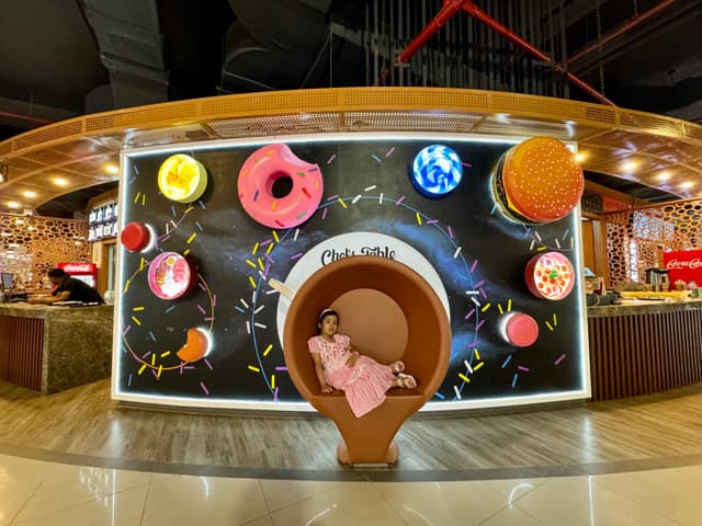 The image depicts an indoor setting with a playful and colorful design theme. There's an illusion of a woman sitting on a toilet paper roll, which is actually a part of the wall decoration featuring a creative arrangement of images and objects that mimic a real-life scenario. This artistic setup includes various items like donuts, planets, and other whimsical elements. The background shows a cafe or restaurant environment with a counter displaying pastries and beverages.