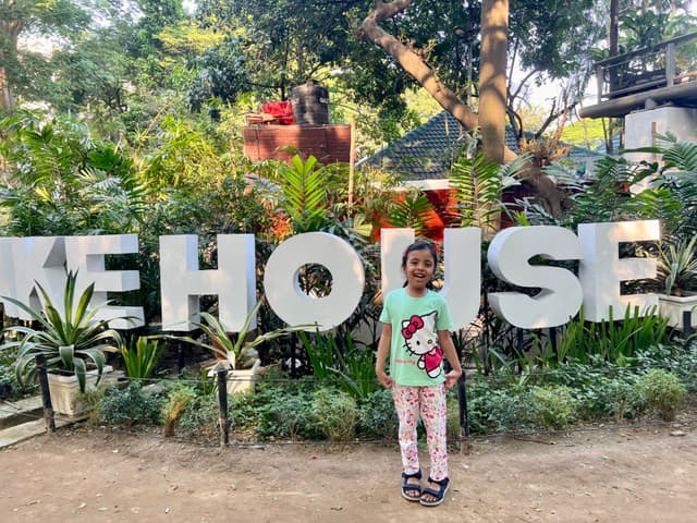 The image depicts a person standing in front of a large sign that reads "SKATEHOUSE." The individual appears to be a child wearing a patterned top and dark pants, with their hair up. They are holding what seems to be a small pink object near their face. The sign is positioned behind the person, suggesting they are posing for a photo. In the background, there are trees and other greenery, indicating an outdoor setting.