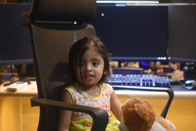 The image shows a young child sitting in an office chair at a desk, with their attention directed towards something off-camera. Behind the child, there's a workspace setup, including a computer monitor and keyboard to the right. The child is wearing casual attire and appears to be engaged in something happening out of frame. There are no texts visible in this image.