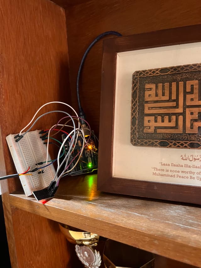 The image appears to show an electronic device, possibly a music or sound player, with various wires and connectors attached to it. The device has a small screen displaying what seems to be Arabic script. It's placed inside a shelf or drawer with a wooden finish. There is a digital readout on the front of the device that displays some kind of data or status information, which is not fully visible due to the angle and perspective of the photo.