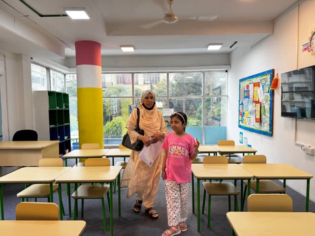 The image depicts an interior space that appears to be a classroom or educational setting. There are desks and chairs arranged for group seating, and the room has a whiteboard at one end. A young child, who seems to be wearing a pink shirt and pants, stands next to a woman with a headscarf. They both appear to be observing something in the distance. The room is well-lit and looks clean and orderly. The walls are decorated with what appears to be colorful educational posters or materials. The environment suggests an educational institution or community center, possibly offering language or other courses given the visual aids on the walls.