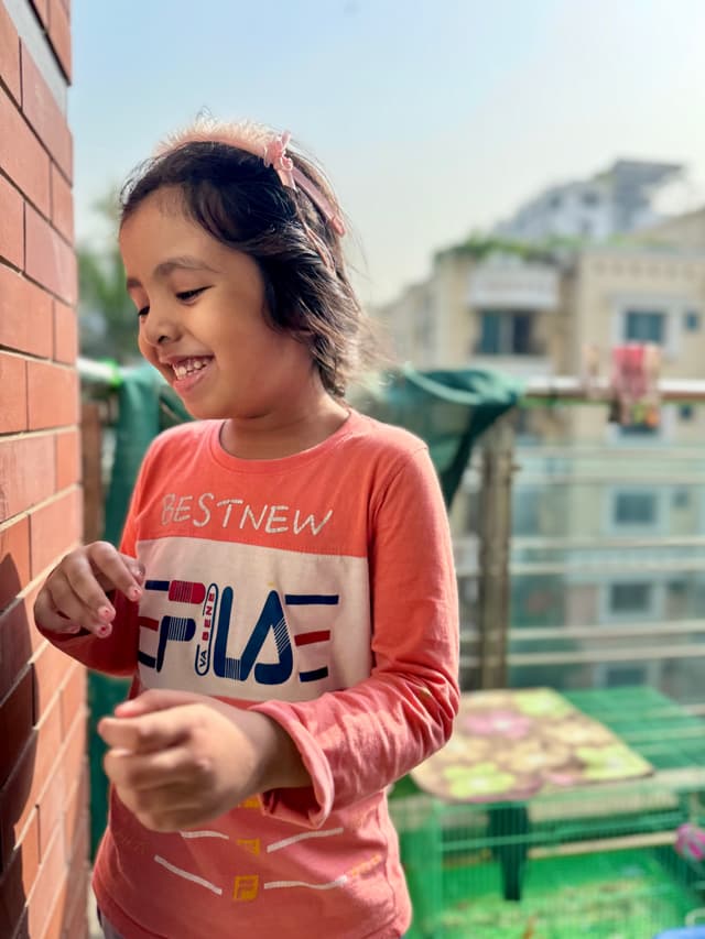 The image features a young girl with dark hair, smiling and looking down at something in her hand. She appears to be on a balcony as suggested by the presence of railings in the background. The balcony has a brick wall and a glass floor panel. The child's attire includes a pink top with what seems to be an emblem or logo on it, and she is wearing glasses. The image conveys a sense of joy and curiosity.