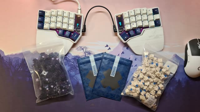 The image displays a desk with various items on it. In the foreground, there is a keyboard with a colorful key layout that resembles a customizable or ergonomic keyboard design. Behind the keyboard, there are two bags filled with what appears to be small, blue pellet-like objects, possibly related to computer peripherals or electronics. These bags have labels indicating they contain "Rice," "Keycaps," and "Mouse." To the right of the desk, there's a collection of keycaps, each corresponding to one of the keys on the keyboard in front. In addition, there are some small items that could be components for assembling or repairing electronic devices. The background features a pink wall with a faint landscape image and a window.
