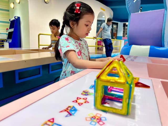 The image depicts an indoor setting, likely a children's play area or a section within a larger building. In the foreground, there is a child seated at a table with various toys scattered around, including what appears to be a large dollhouse with colorful blocks and a small toy house on the table in front of the child. The child seems engaged in playing with the toys. Behind the child, there are other people present, though they are not the focus of this image.

The environment suggests it could be an educational or entertainment space designed for young children, given the presence of a play area and the variety of toys. The design is modern and colorful, aimed at attracting and engaging young visitors.