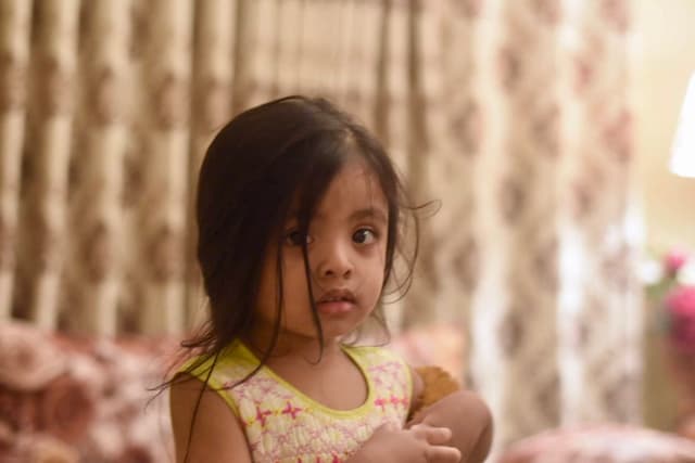 The image displays a young child looking directly at the camera. The child has dark hair and appears to be indoors, with a floral wall behind them. Their expression is neutral, and they seem to be seated on what could be a couch or chair. There's no additional context or items in the scene that provide further details about the location or setting.