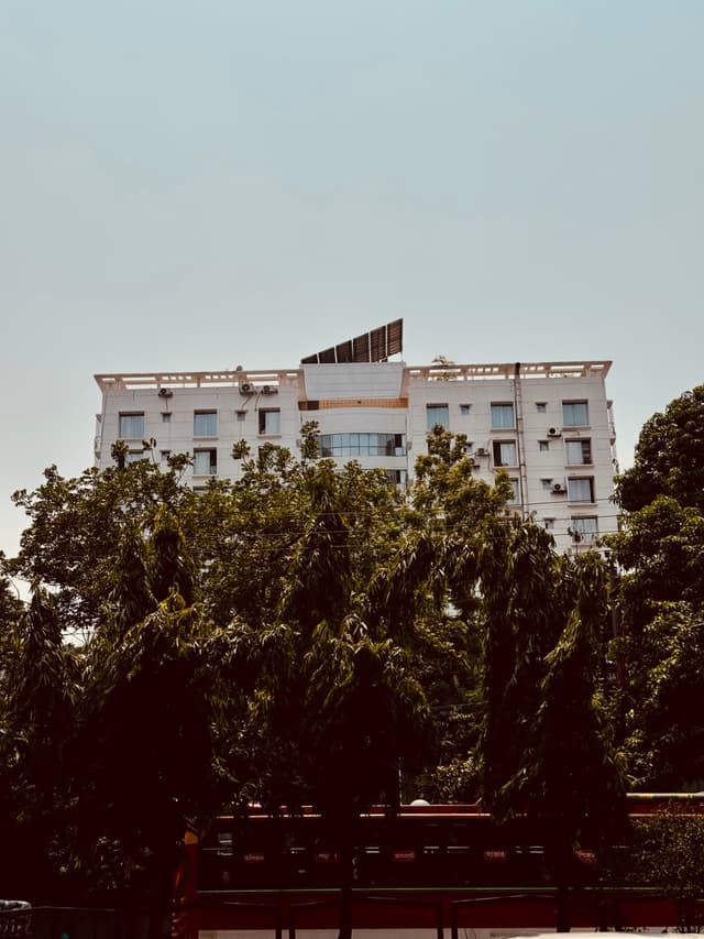 The image displays a multi-story building with a visible balcony. The photograph has an overlay effect, causing parts of the structure to appear fragmented and distorted, creating a pixelated appearance. The background features a clear sky and lush greenery, which indicates that the building is surrounded by trees or plants. There's also a partial view of a power line. The style of the image appears to be digitally manipulated to create an artistic effect with the overlay.