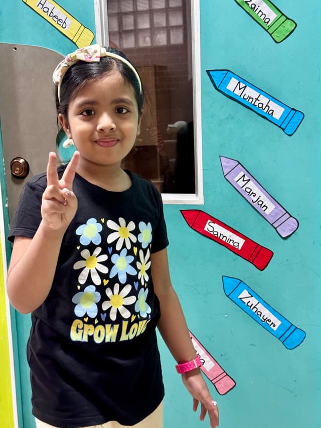In the image, there's a young girl who appears to be posing for the photo. She has short hair and is wearing what seems to be a black t-shirt with some sort of design on it. The background is indoors with a colorful wall that has a vertical row of what looks like crayons or colored pencils in various colors, all pointing upwards. There's a window on the upper part of the wall. To the left of the girl, there's a door partially visible with some text on it, but it's not clear enough to read. The overall atmosphere is casual and cheerful.