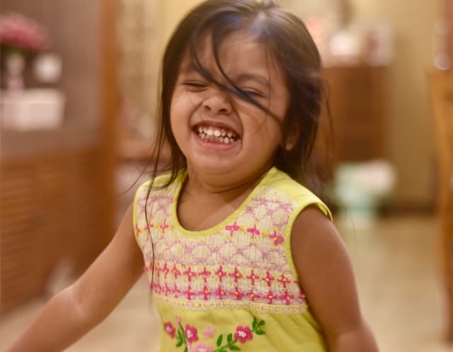 The image shows a young girl with her mouth open, laughing and displaying joy. She has dark hair and is wearing a light-colored sleeveless top with a pattern on it. The background is blurred but appears to be an indoor setting with a cabinet and a counter visible.