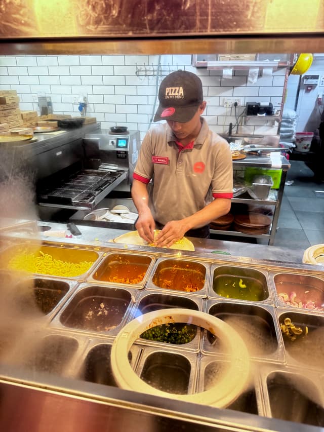 This image depicts a restaurant kitchen scene. A chef, wearing a white apron and a black cap with red lettering, is working at a counter in front of open food containers and various condiments. The chef appears to be preparing food, focusing on the task at hand. In the background, there are additional counter spaces equipped with more cooking tools and condiments. The image captures the bustling atmosphere of a commercial kitchen.