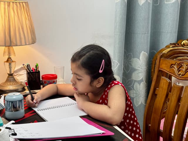 The image shows a young girl sitting at a desk in front of an open laptop, actively engaged in some task. She appears to be at her own study area with various items like a notepad and a book nearby.